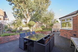 Een terras met een tafel met glazen blad, omringd door rieten stoelen, een bakstenen muur en een houten omheining.