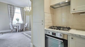 A modern kitchen with a gas stove, oven, and stainless steel range hood. There is a dining area with a table and chairs visible through an open door.