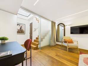 A modern living room with a wooden floor, a dining table, a red chair, a sofa, a TV, and a staircase leading to another room.