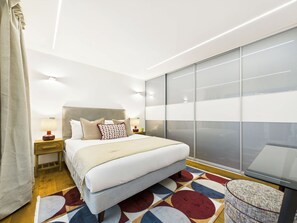 A modern bedroom with a large bed, a bedside table with a lamp, a patterned rug, and a large window with blinds. - Stylish & Designer Camden Town Apartment (Greater London)