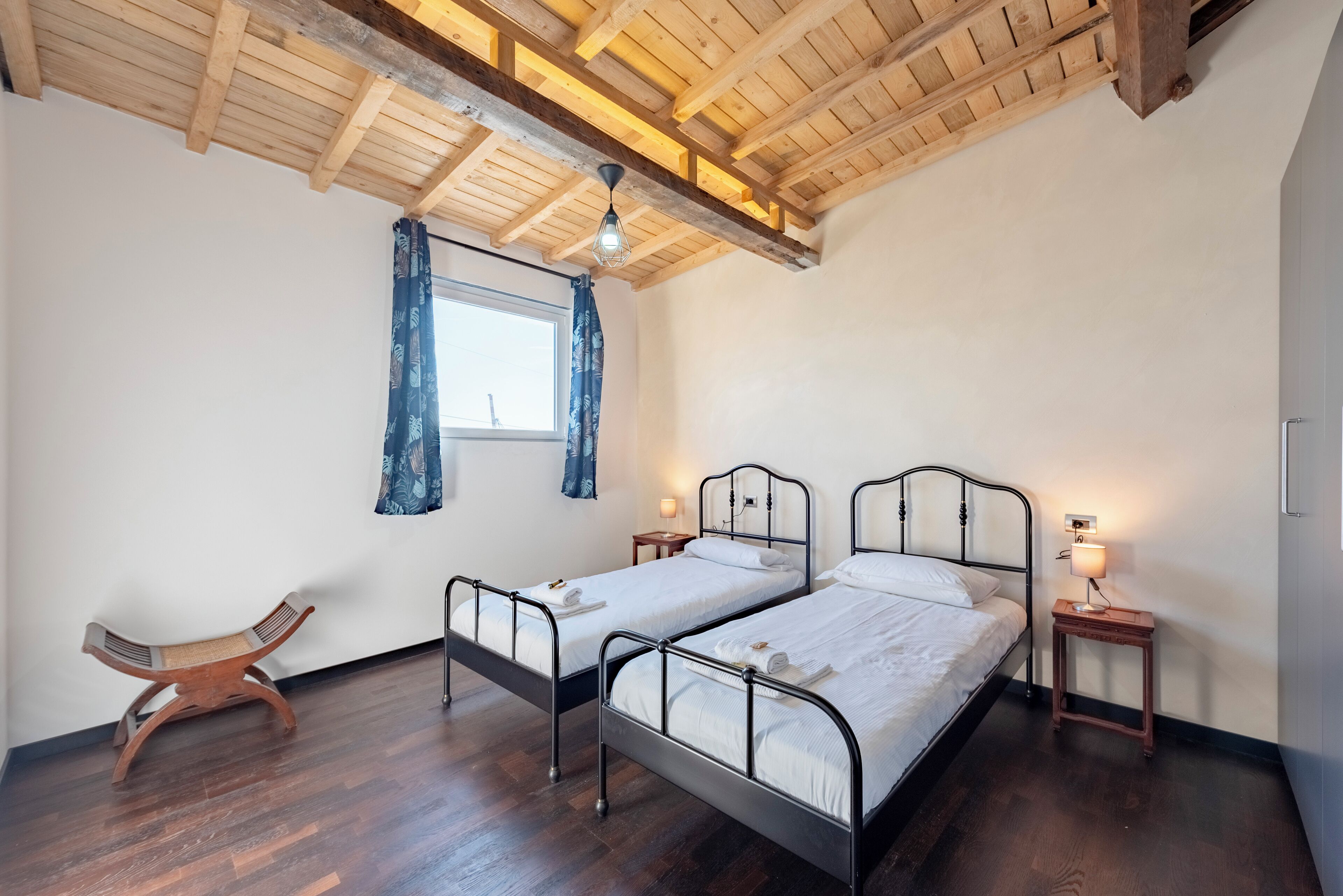 Quarto com duas camas de metal, piso de madeira, uma cadeira de madeira, janela com cortinas azuis e luminária pendente.