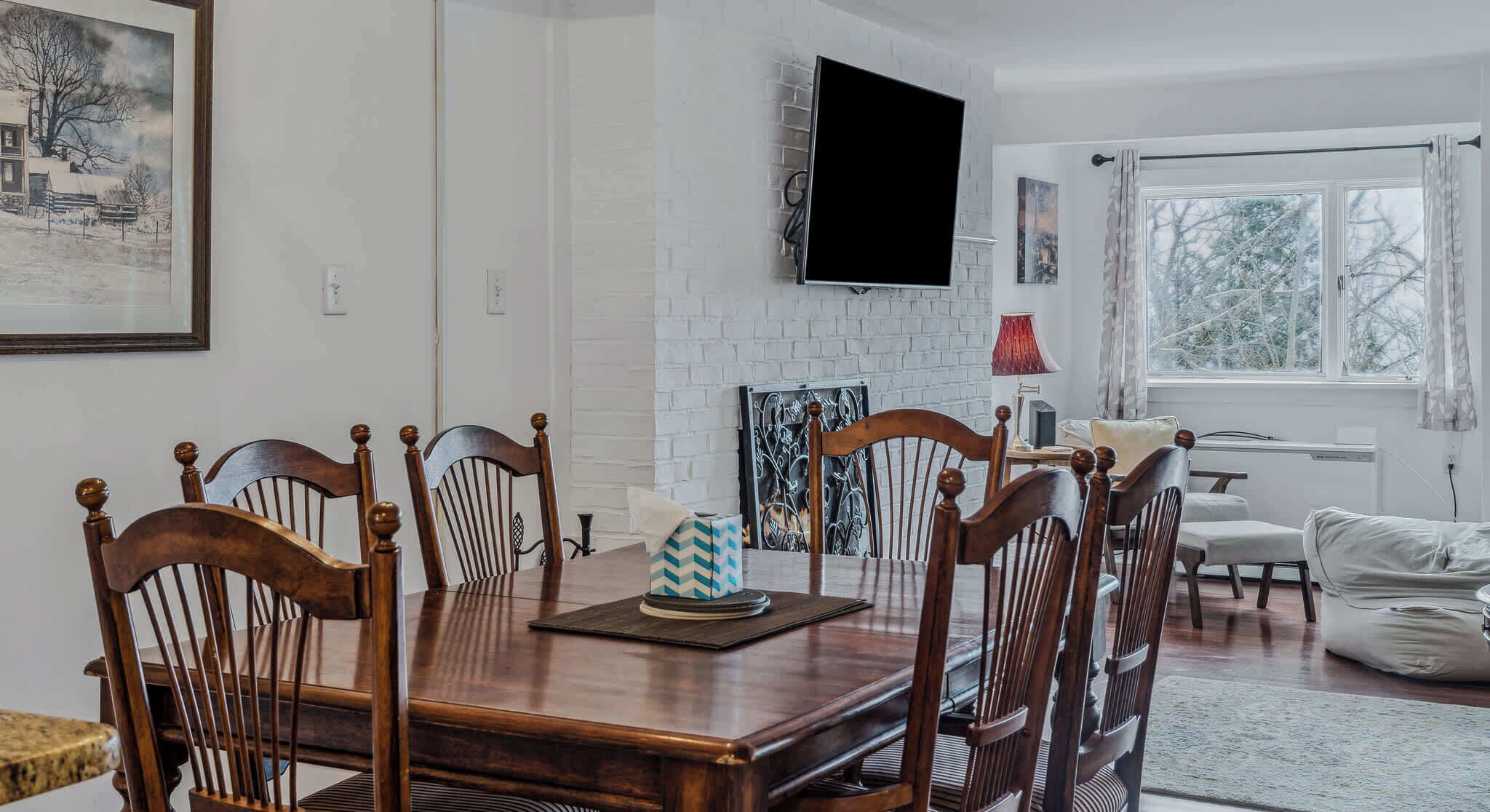 A dining area with a wooden table and chairs, a fireplace, a flat-screen TV, and a window with a view of trees.