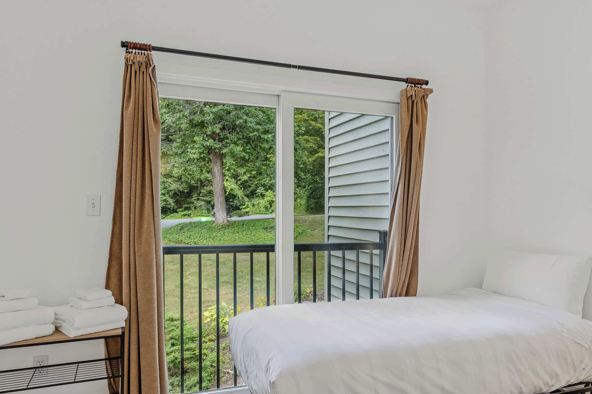 Una camera da letto con un letto, un tavolino con asciugamani, un balcone con tende e vista sul verde.