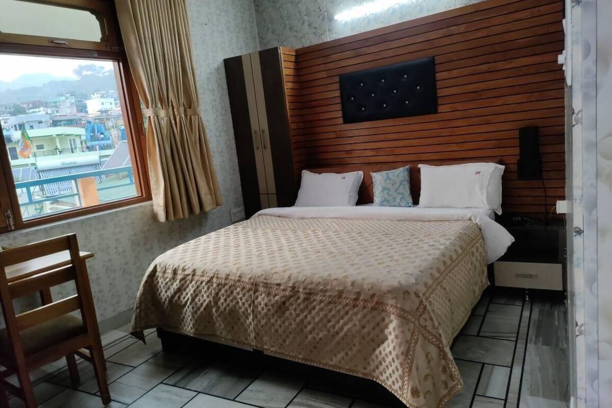 Un dormitorio con cama, cabecero de madera, mesita de noche, silla y ventana con cortinas.