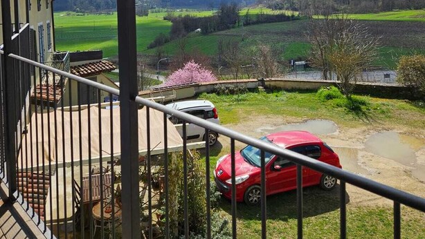 Un balcon donnant sur une voiture rouge garée sur une pelouse, une pergola en bois au-dessus et un paysage rural en arrière-plan.