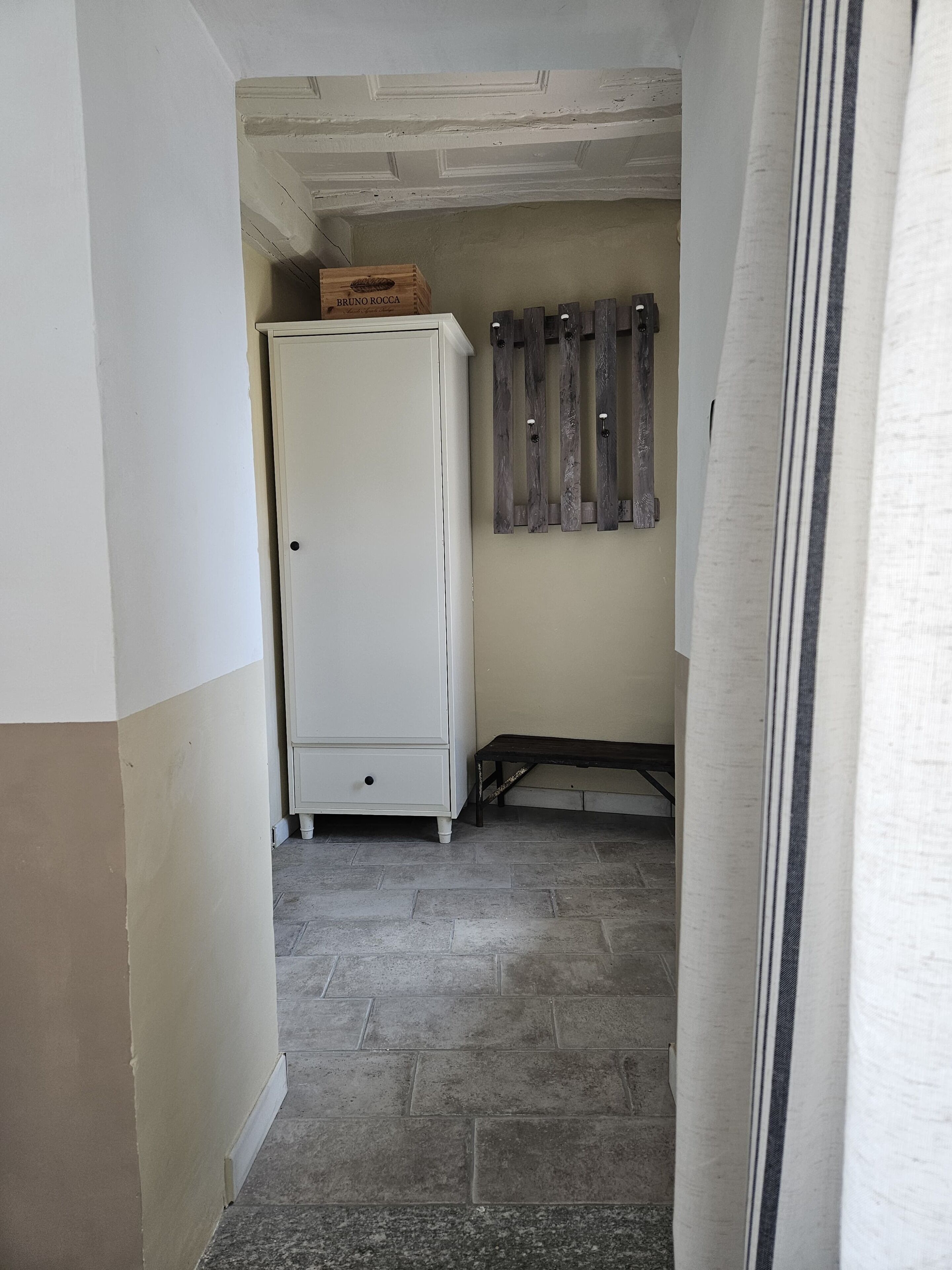 Un couloir étroit avec une armoire blanche, une caisse en bois portant l’inscription « BRUNO ROCCA » et une étagère en bois fixée au mur.