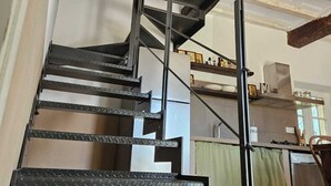 Un escalier en colimaçon avec des rampes en métal et une finition en bois, menant à un espace cuisine équipé d’un plan de travail et d’appareils électroménagers.