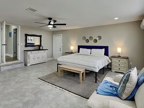 A bedroom with a bed, bedside tables, a bench, a ceiling fan, a mirror, and a door.
