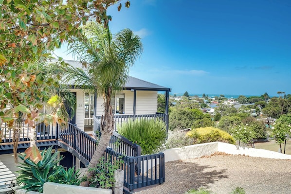 A two-story house with a balcony, surrounded by greenery and overlooking a coastal view.