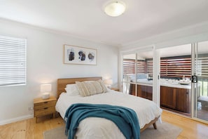A bedroom with a bed, bedside tables, a nightstand, a window with blinds, a ceiling light, and a sliding glass door leading to a balcony with a view.