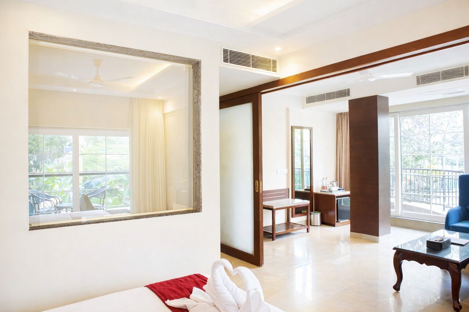 A hotel room with a large mirror, a bed with a red blanket, a wooden table, and a view of the outdoors.