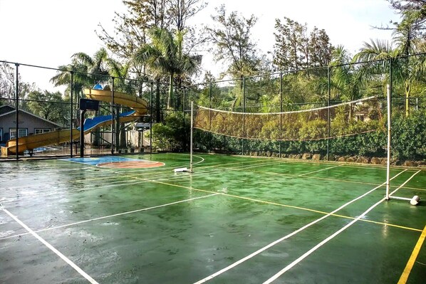 Un court de tennis en plein air, doté d’un filet, entouré d’arbres et agrémenté d’un toboggan aquatique.
