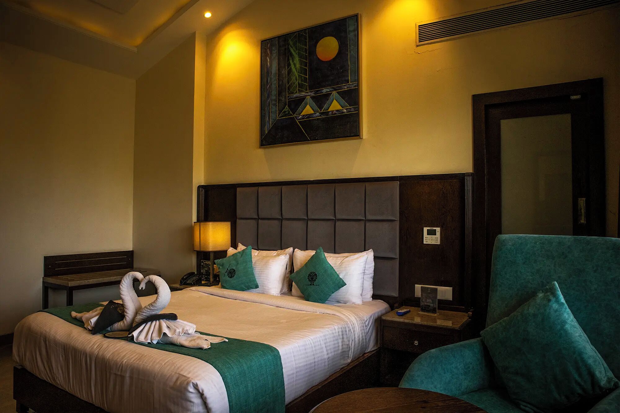 Une chambre d’hôtel avec un grand lit, une tête de lit, une table de chevet, une lampe, une horloge murale, un miroir et un fauteuil vert.