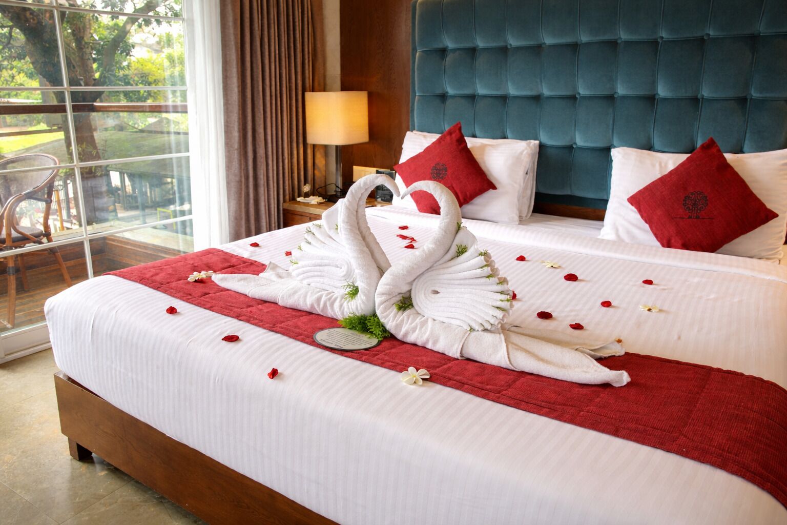 A hotel room with a bed featuring a swan towel arrangement, red pillows, and a view through a large window.