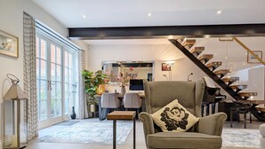 A modern living room with a glass coffee table, a comfortable armchair with a decorative pillow, a dining area with a table and chairs, and a staircase leading to an upper level.