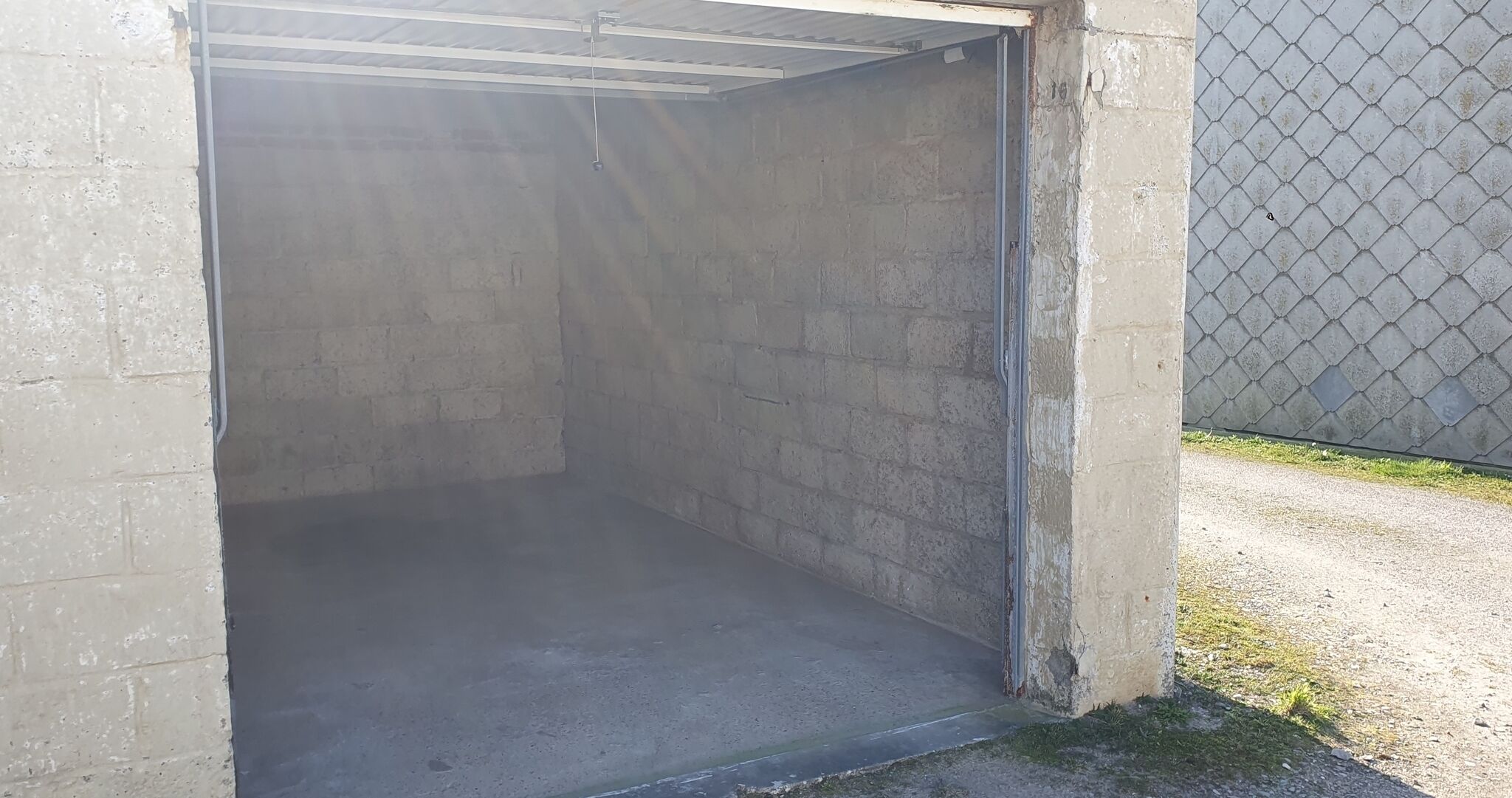 Eine Garagentür aus Beton mit Metallrahmen, ein Betonboden und ein Zaun aus Maschendraht im Hintergrund.