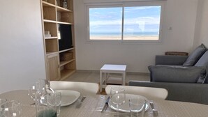 A dining area with a table set for four, a bookshelf, a grey sofa, and a window with a beach view. - 🌊 Fenêtres sur Mer - Sea View & Garage - 3 bedrooms (Berck)