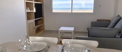 A dining area with a table set for four, a bookshelf, a grey sofa, and a window with a beach view.