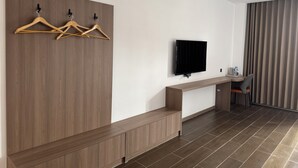 A modern hotel room with a wooden panel wall, a flat-screen TV mounted above a wooden console table, a seating area with a chair, and a large window with curtains. - Xianglong Hotel (Luang Prabang)
