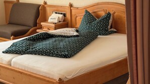 A wooden bed with a geometric-patterned blanket, a bedside table with a towel, and a chair in a room with wooden paneling and a wall-mounted lamp. - In & Out boarding house comfort room (Stolberg)