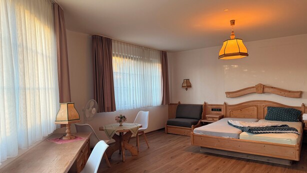 A hotel room with a wooden bed, a desk with a chair, a small table with a vase of flowers, a sofa, and a ceiling lamp. - In & Out boarding house comfort room (Stolberg)
