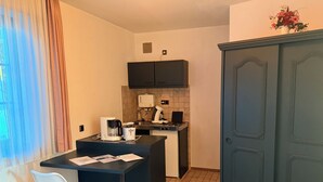 A compact kitchen with a coffee maker, a kettle, and a small stove. There is a white chair and a dark blue cabinet. - In & Out boarding house comfort room (Stolberg)