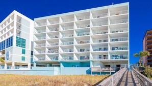 A multi-story hotel with a white and blue facade, multiple balconies, and a boardwalk leading to the entrance. - Emerald Skye 63 1 Bedroom Condo (Orange Beach)