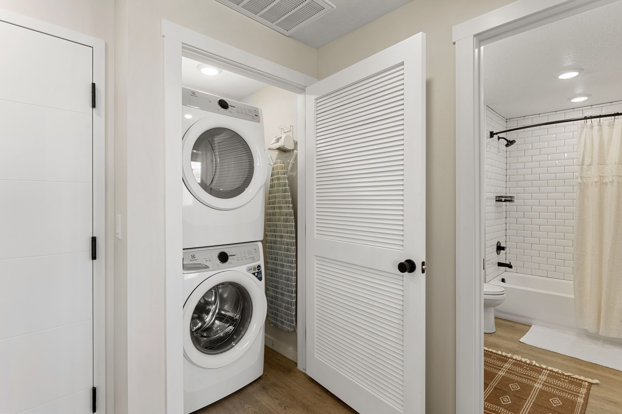A laundry room with a front-loading washer and dryer, a towel rack, and a shower area with a white tiled wall and a beige shower curtain.