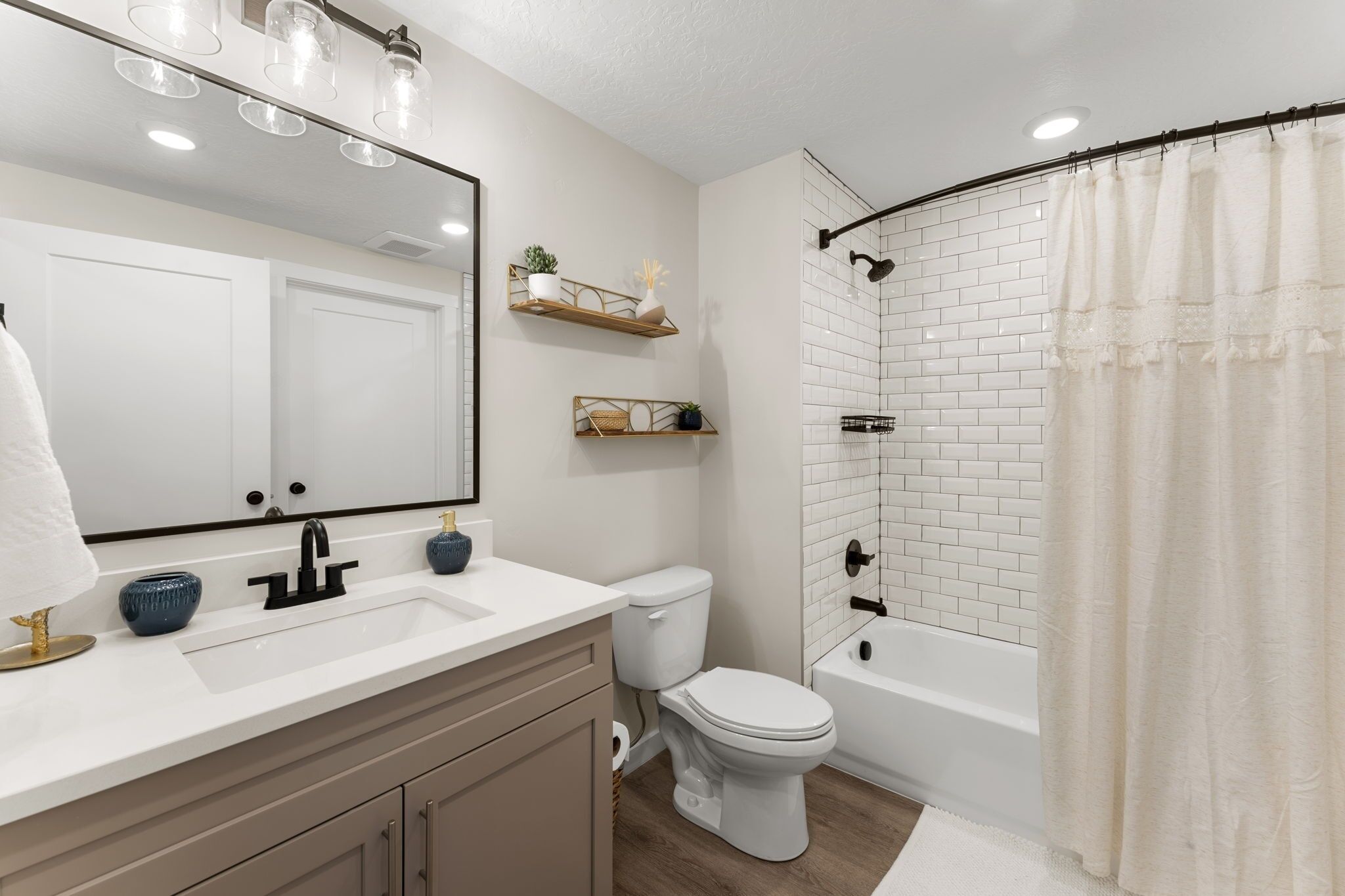 A modern bathroom with a white sink, toilet, and bathtub, featuring a white tiled shower area and wooden shelves with decorative items.
