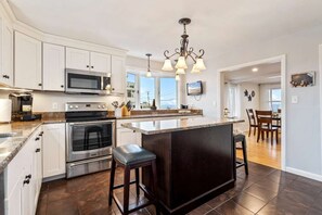 Una cocina moderna con armarios blancos, electrodomésticos de acero inoxidable, una isla grande con taburetes y un área de comedor visible a través de una puerta abierta.