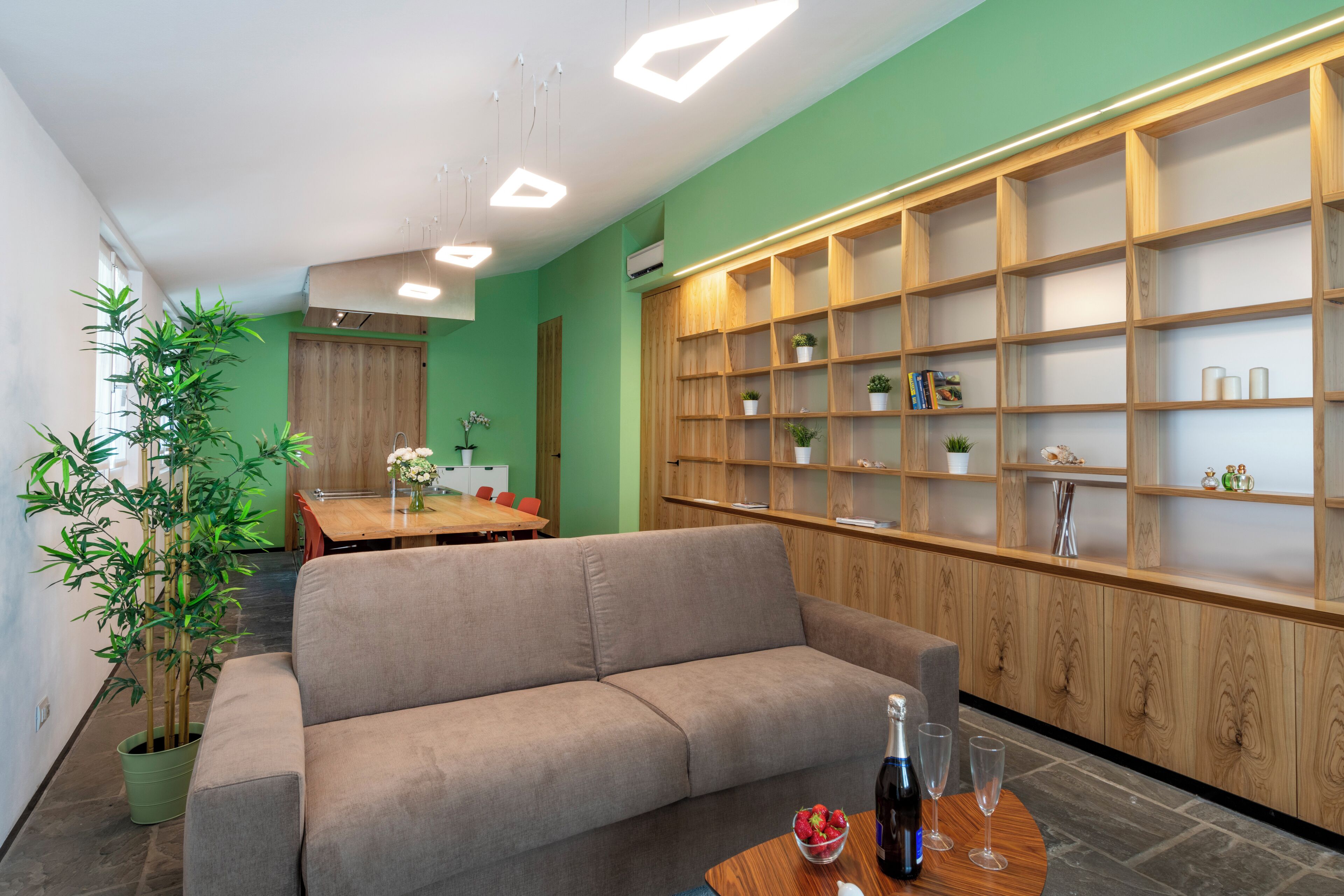 A modern living room with a beige sofa, a wooden bookshelf, and a dining area in the background.