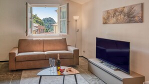 A living room with a sofa, a flat-screen TV, a table with a bottle and glasses, and a window with a view of buildings and trees. - Charming Portofino 3-bed retreat (Portofino)