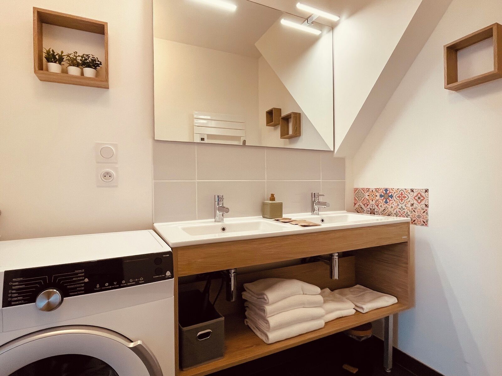 A modern bathroom with a dual sink, a washing machine, and a wooden shelf with towels.