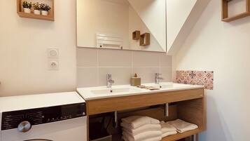 A modern bathroom with a dual sink, a washing machine, and a wooden shelf with towels.