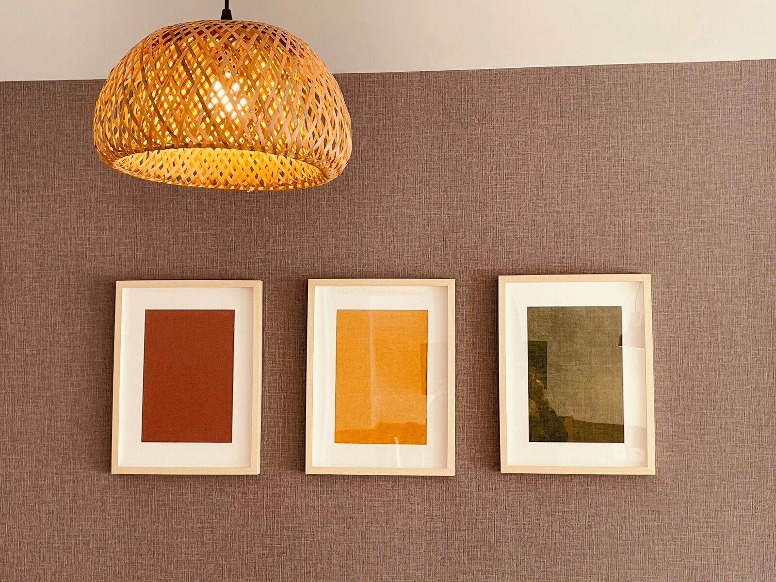 Ein Zimmer mit einer gewebten Pendelleuchte, drei gerahmten Kunstwerken an einer strukturierten Wand und einem neutral gefärbten Sofa.