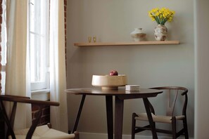 Un coin repas avec une table ronde, deux chaises, une étagère en bois au-dessus et une fenêtre avec des rideaux.