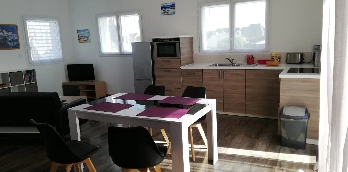 A modern kitchen with a built-in oven, a sink, and a coffee machine. There is a dining area with a table and chairs.