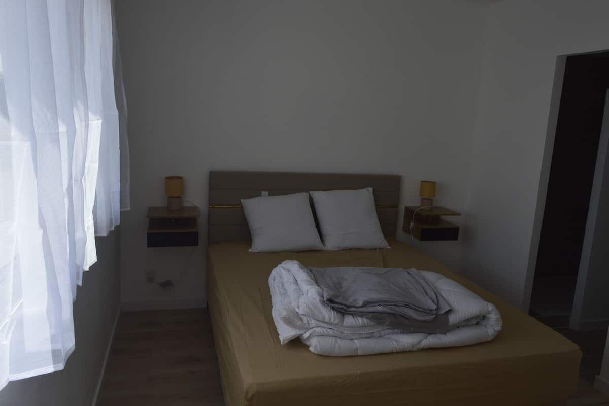 A bedroom with a bed, two bedside tables, a headboard, and a window with sheer curtains.