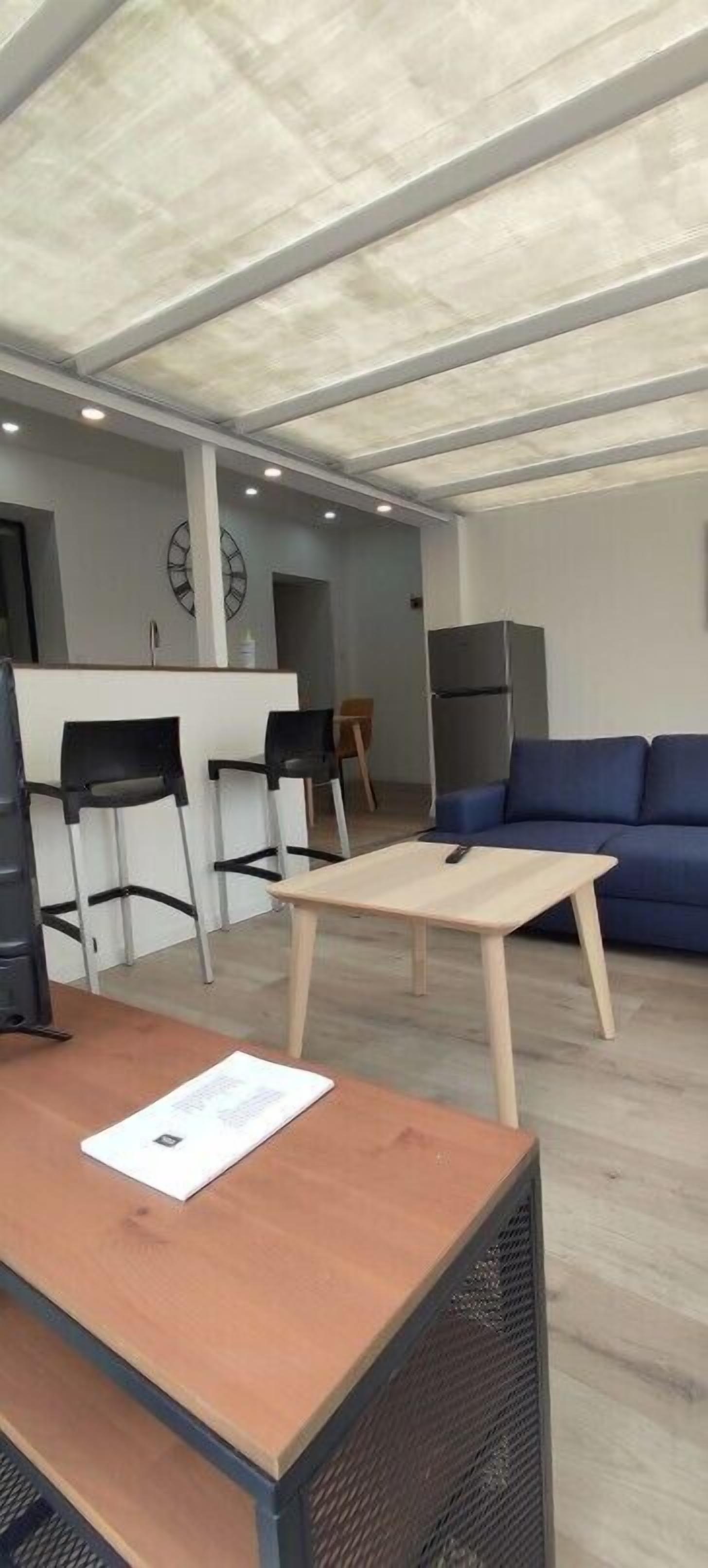 Een moderne woonkamer met een houten salontafel, een blauwe bank en een klein keukenhoekje met een koelkast en stoelen.