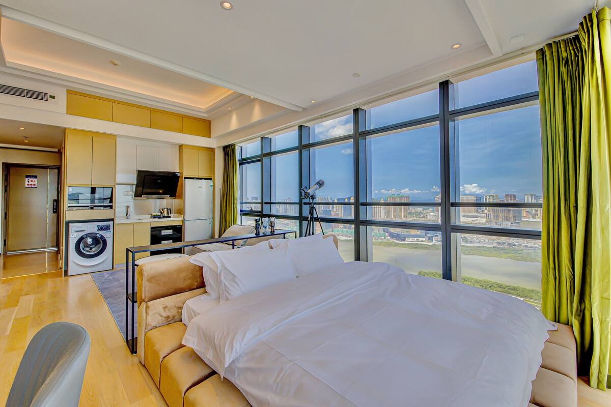 Una habitación de hotel moderna con una cama grande, kitchenette, lavadora y vistas de la ciudad.