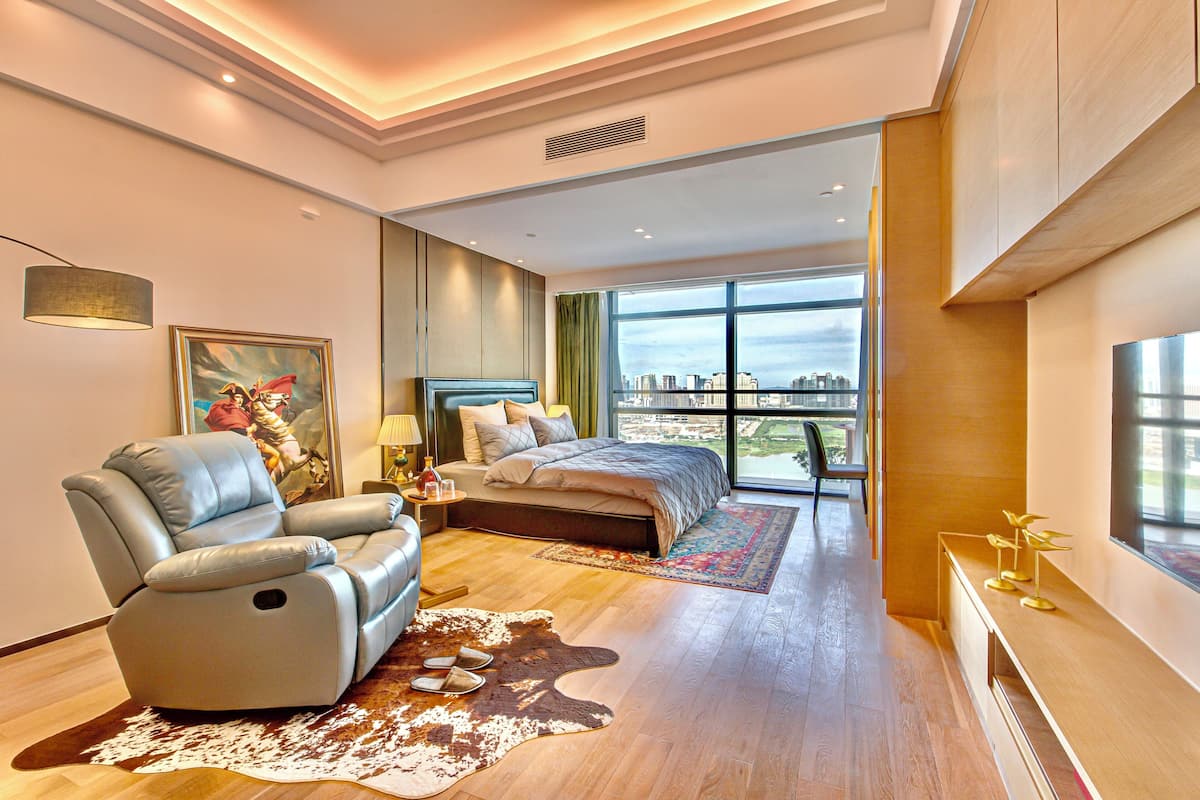 Una habitación de hotel moderna con una cama grande, un sillón de cuero, vistas a la ciudad y suelo de madera.
