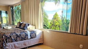 Ein Schlafzimmer mit großem Fenster, einem Bett mit gemusterten Betttextilien, einem schwarzen Teppich und einer Deckenleuchte.