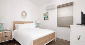 A bedroom with a bed, bedside tables, a wall-mounted air conditioner, a window with blinds, and a small desk with a television.