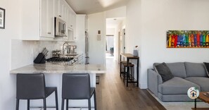 A modern kitchen with white cabinets, a marble countertop, and stainless steel appliances. There is a dining area with a gray sofa and a colorful artwork on the wall.