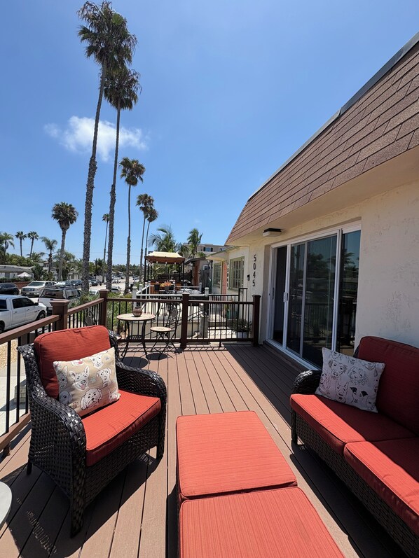 A deck with wicker furniture, a red cushion, and a view of palm trees and parked cars.