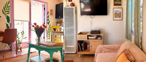 Un saló amb un sofà, una televisió penjada a la paret, una taula amb un ram de flors i un aparell d'aire condicionat alt.