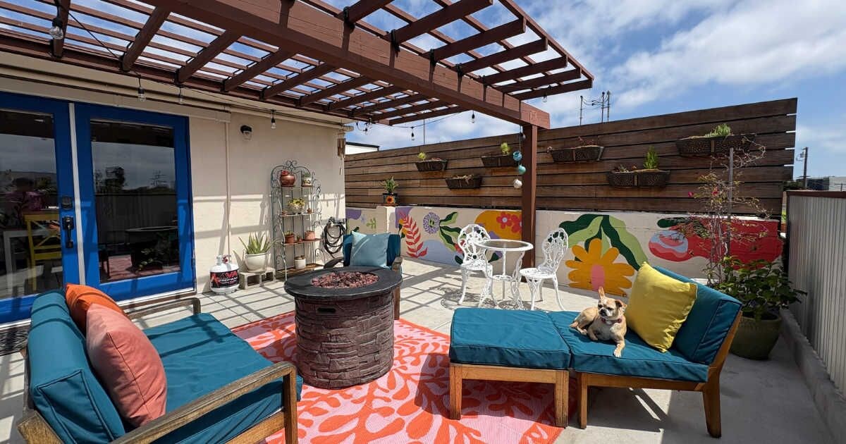 Een terras buiten met een houten pergola, een zithoek met kussens, een ronde tafel en een hond.