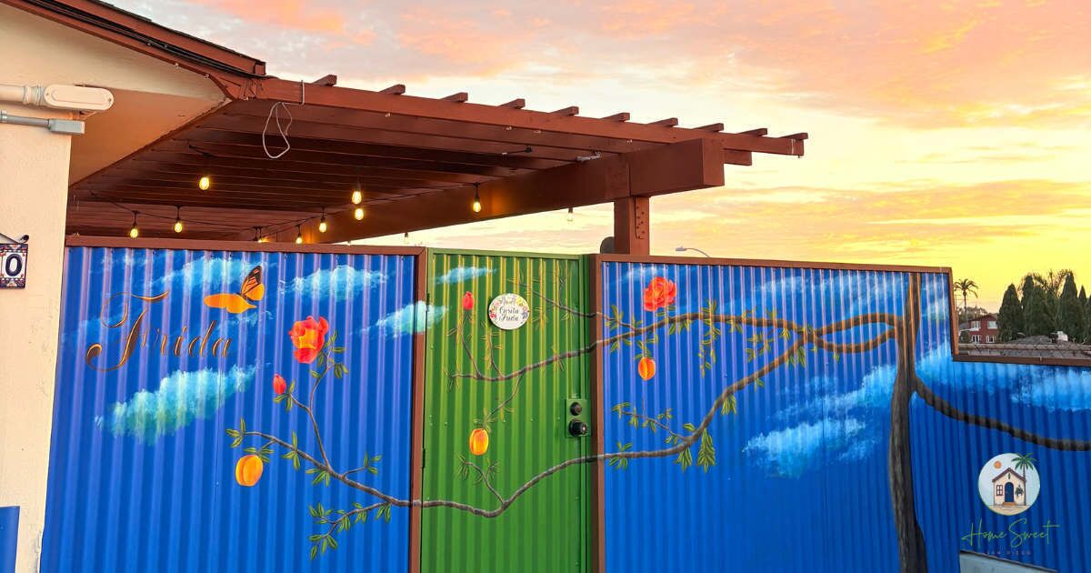 Cerca de metal corrugado com ramos e frutos pintados, uma porta verde e uma pérgula com luzes.