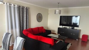 Un salon comprenant un canapé d’angle, un téléviseur, une horloge et un coin repas avec des chaises et une table.
