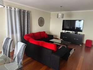 A living room with a sectional sofa, a TV, a clock, and a dining area with chairs and a table.
